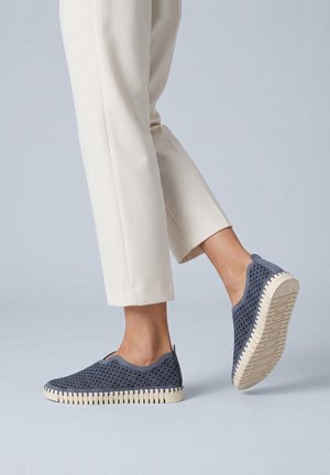 Persona que lleva pantalones beige claro y zapatillas tipo slip-on azul oscuro con suelas texturizadas, un pie levantado mostrando el patrón de la suela del zapato.