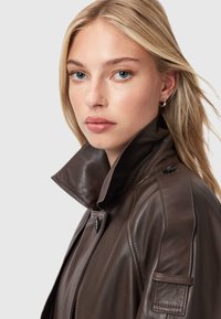 Bruine leren jas met een hoge kraag, slank ontwerp en zichtbare knoopsluiting. Heeft een gestructureerd oppervlak en schouderdetails.