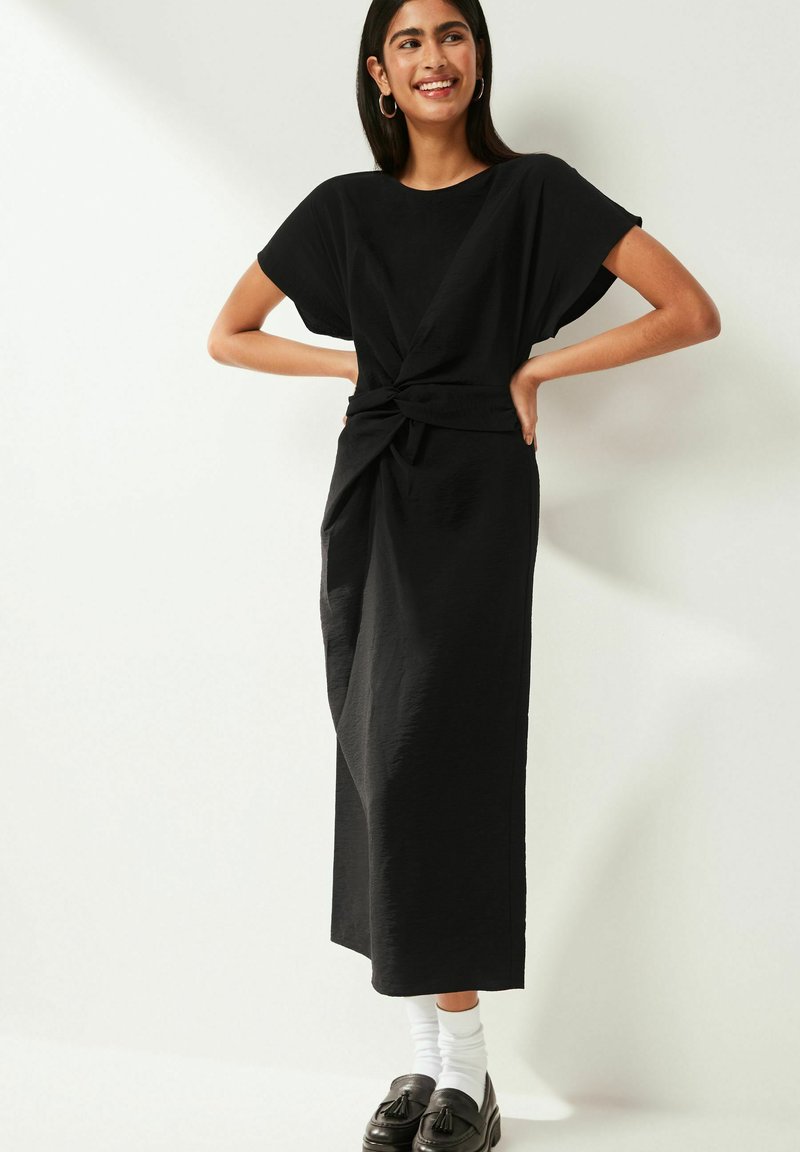 Donna sorridente che indossa un vestito midi nero con dettaglio a nodo frontale, calze bianche e mocassini neri con frange, in piedi contro uno sfondo bianco.