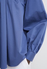 Personne à la peau foncée portant une chemise bleue à manches longues amples avec des manches bouffantes et un pantalon blanc, main dans la poche.