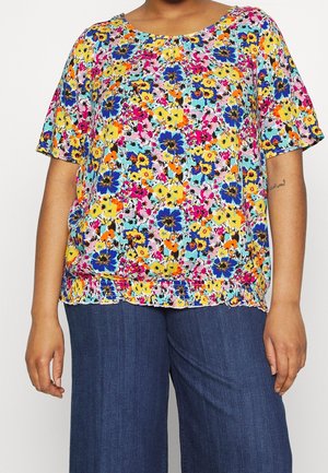 Frau trägt eine kurzärmelige, mehrfarbige Blumenbluse mit elastischem Saum und dunkelblaue, weite Hosen.