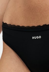 Gros plan sur un sous-vêtement noir avec bordure en dentelle et le nom de la marque HUGO imprimé en blanc sur le tissu contre la peau.