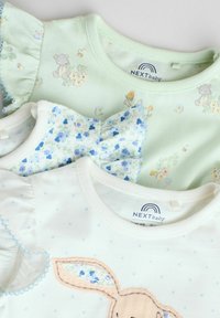 Tre babyt-shirts lagd oven på hinanden: mintgrøn med blomsterprint, lyseblå med blomster og flæser, samt hvid med polka prikker og kaninbroderi.