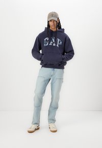 Jūras spēku kapuce ar "GAP" logo raibās krāsās, saskaņota ar gaiši ziliem džinsiem un baltām kedām. Modelis valkā gaiši pelēku cepuri.