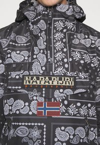 Zwarte jas met patroon en witte paisley ontwerpen, rits borstzak, Napapijri logo patch en Noorse vlag patch aan de voorkant.