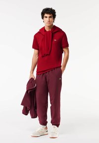 Bordeaux polo skjorte med en sweatshirt draperet om halsen, matchende joggers og hvide sneakers med orange detaljer. Glatte teksturer overalt.