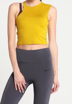 Femme portant un crop top sans manches jaune avec une bretelle de soutien-gorge marron visible et des leggings taille haute gris foncé avec un petit logo.