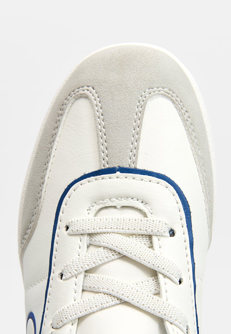 Baskets blanches avec des accents bleus, présentant un capuchon en suède texturé et un corps en cuir lisse. Les lacets sont gris clair, avec des détails de couture.