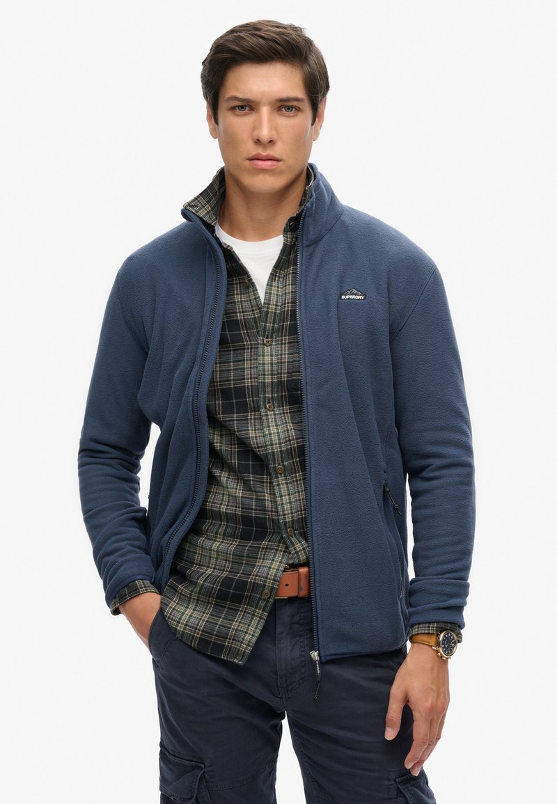 Granatowa polarowa kurtka z zamkiem z przodu, noszona na ciemną koszulę w kratę. Posiada małe logo na piersi i jest zestawiona z granatowymi spodniami cargo.