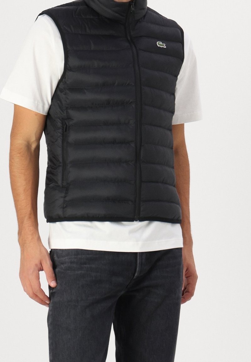 Homme portant un gilet matelassé noir sans manches sur un t-shirt blanc à manches courtes et un jean foncé devant un fond uni.