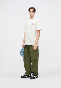 Κρεμ βαμβακερό μ�πλουζάκι με ροζ γραφικό σχέδιο, σε συνδυασμό με ελαιόχρωμο παντελόνι cargo με πολλές τσέπες και μαύρα αθλητικά παπούτσια.