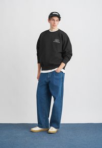 Črna trenerka z belim logotipom, široke modre kavbojke in bele čevlje. Kapa je črna z belim logotipom. Navaden bel majica je vidna.