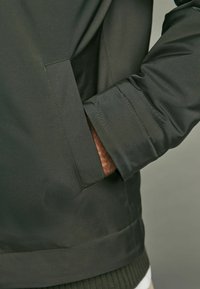 Mørkegrøn jakke med en glat tekstur, der har en sidelomme og ribbede manchetter. Syet detaljering langs sømmene.