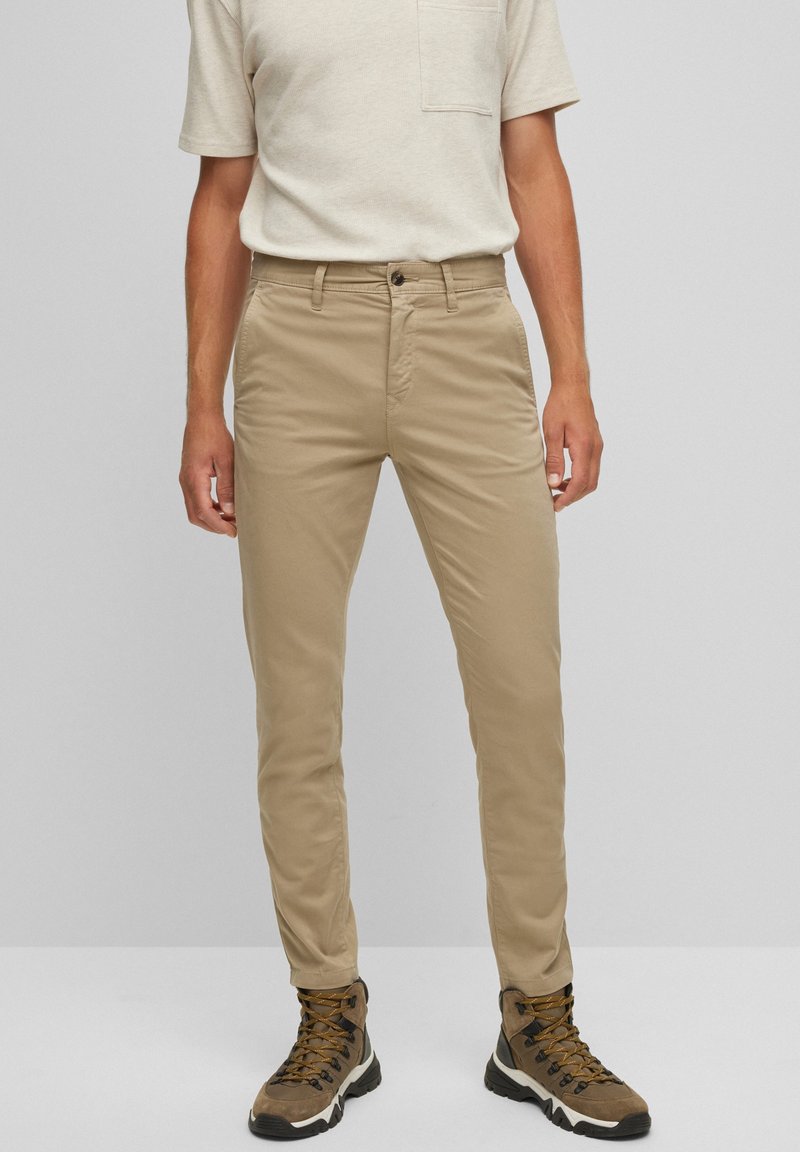 Man i beiga slim-fit byxor, ljusbeige kortärmad skjorta och bruna vandringsskor, står mot en enfärgad grå bakgrund.