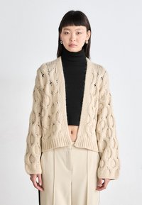 Πλεχτό ανοιχτό κασκόλ σε μπεζ, με υφή και ανοιχτό σχέδιο, διαθέτει χαλαρή εφαρμογή και κοντό μήκος. Συνδυάζεται με μαύρο λαιμόκοψη.