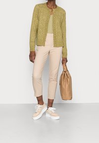 Woman wearing a green knitted cardigan, beige cropped pants, cream sneakers, and holding a tan handbag against a plain background.