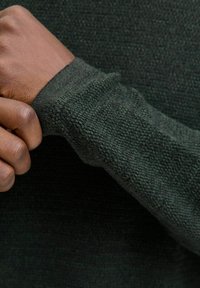 Szczegół ciemnozielonego swetra robionego na drutach, ukazujący teksturowany materiał oraz mankiet trzymany w dłoni, podkreślający miękkość i wzór tkaniny.