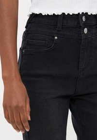 Mustat farkut ovat korkea vyötärö, niissä on kaksi etutaskua ja hopeinen nappisulku. Kangas on sileä ja hieman tekstuuri.