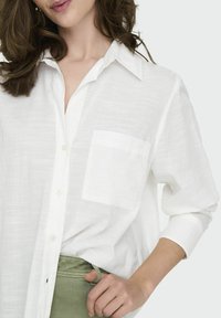 White button-up shirt with a relaxed fit, collar, and a single chest pocket, made from lightweight, textured fabric. Paired with olive green pants.