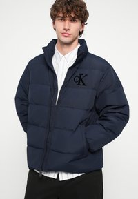 Námornícka modrá prešívaná bunda, zapínanie na zips vpredu, elastické manžety, logo vyšívané na hrudi, s jemným, textúrovaným povrchom a vysokým golierom.