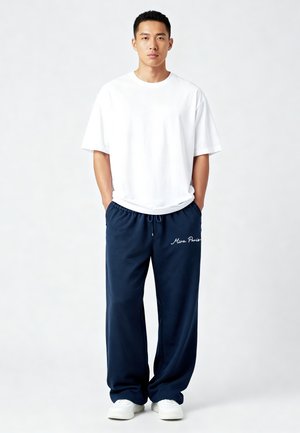 Man staat met handen in de zakken, draagt een wit oversized T-shirt, donkerblauwe broek met de tekst "Mira Paris" en witte sneakers op een effen achtergrond.