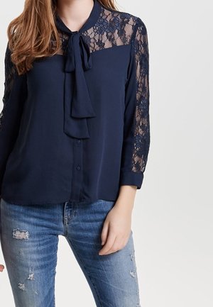 Vrouw die een marineblauwe blouse draagt met kanten mouwen en bovenborst, gecombineerd met versleten blauwe jeans, staand tegen een egale achtergrond.