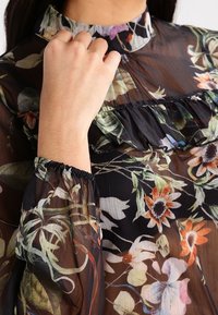 Blouse noire transparente avec un motif floral, encolure froncée et poignets élastiques, présentant des couleurs vives et une texture légère.