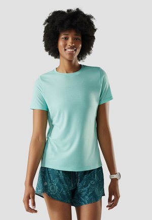 Uśmiechnięta kobieta o kręconych włosach, ubrana w jasnoturkusową koszulkę z krótkim rękawem, ciemne szorty w kwiatowy wzór oraz biały zegarek na nadgarstku.