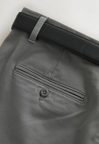 Pantalon gris avec une poche passepoilée boutonnée à l'arrière et une ceinture en cuir noire passant à travers une boucle sur un fond clair.
