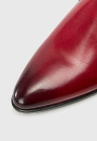 Červená kožená bota s lesklým povrchem, s špičatou špičkou a hladkým texturem. Okraj je jemně zakulacený, což zdůrazňuje detaily řemeslného zpracování.
