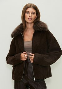 Chaqueta marrón con cremallera y cuello de piel, con una textura suave y un corte holgado. El modelo lleva una blusa beige y pantalones oscuros.