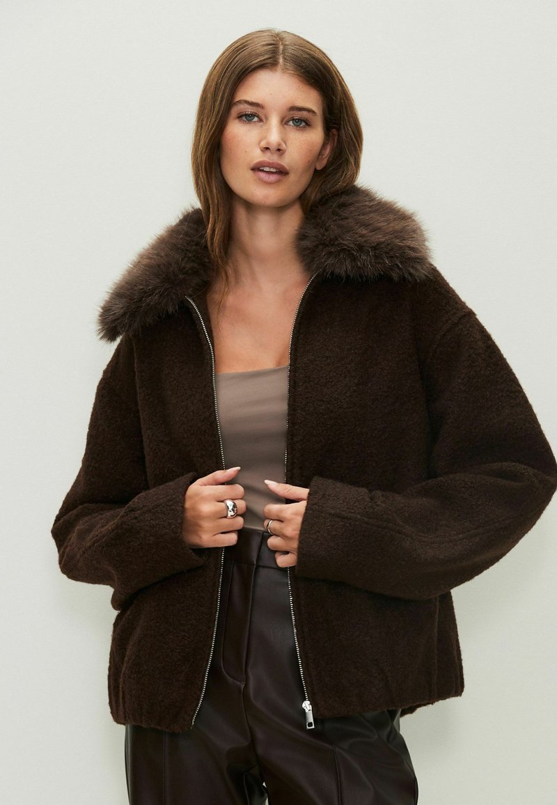 Chaqueta marrón con cremallera y cuello de piel, con una textura suave y un corte holgado. El modelo lleva una blusa beige y pantalones oscuros.