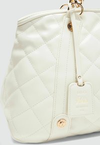 Sac à main en simili cuir blanc matelassé avec des accessoires en or, une étiquette décorative et des accents cousus. Présente une forme arrondie et une fermeture éclair.