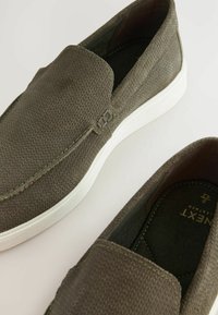 Maslinasto zelene slip-on cipele od teksturiranog materijala, s bijelim gumiranim potplatom i suptilnim detaljima šivanja. Nema vidljivih vezica ili metalnih akcenata.