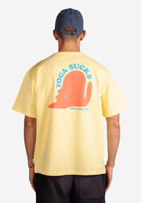 Žluté bavlněné tričko s velkým červeným potiskem na zádech s textem "YOGA SUCKS" a postavičkou v karikaturním stylu. Obsahuje značku.