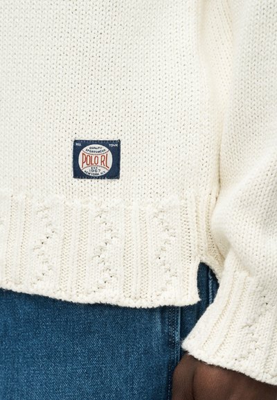 Κρεμ πουλόβερ από πλεκτό ύφασμα με ετικέτα Polo RL σε ναυτικό μπλε και λευκό πάνω από τη ζώνη, φορεμένο με μπλε τζιν και ορατό χέρι με σκουρόχρωμο δέρμα.