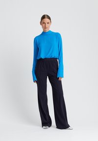 Frau steht vor schlichter Hintergrund, trägt schwarze Hosen mit weitem Bein, ein leuchtend blaues Langarmshirt mit verlängerten Manschetten und silberne spitze Schuhe.