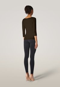 Frau mit lockigem Haar in einem braunen, figurbetonten Oberteil, schwarzen Leggings und nudefarbenen High Heels, die mit dem Rücken zu einer schlichten Hintergrund steht.