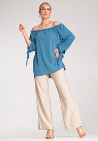 Top w kolorze turkusowym bez ramiączek z długimi rękawami i detalami w postaci wiązań, zestawiony z szerokimi spodniami w kolorze kremowym i przezroczystymi sandałami na obcasie.