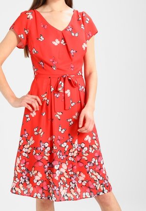 Femme portant une robe rouge longueur genou avec un imprimé papillon et une ceinture nouée à la taille, debout, une main sur la hanche.