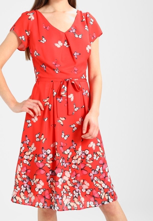 Femme portant une robe rouge longueur genou avec un imprimé papillon et une ceinture nouée à la taille, debout, une main sur la hanche.