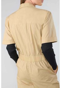 Salopette kaki avec des manches courtes, une taille élastique et une texture lisse. Des manches en ribbed noires sont visibles.