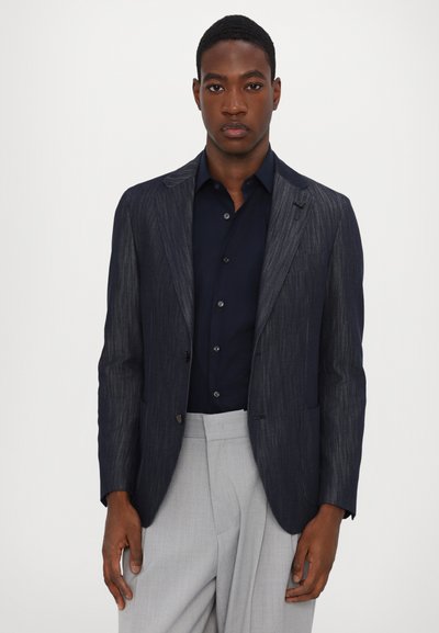 Dunkel gestreifter, taillierter Blazer mit Reverskragen, vorderem Knopf und aufgesetzten Taschen, kombiniert mit einem marineblauen Hemd und hellgrauen, taillierten Hosen.