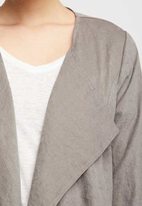 Chaqueta de un gris claro y textura, con un cierre abierto, que presenta un ligero drapeado y una camisa blanca de cuello en V debajo. Tejido suave con un corte relajado.