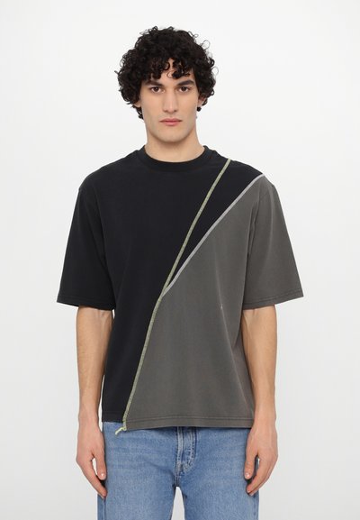 T-shirt bloccato nei colori nero e grigio con maniche corte, caratterizzato da una cucitura diagonale accentuata da cuciture verdi. Materiale in cotone testurizzato.