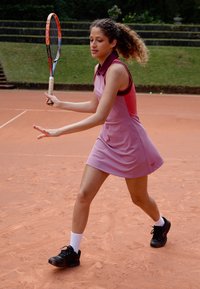 Tennis dress in light pink with a dark pink collar and side panels. Black athletic shoes and white socks, on a clay court background.