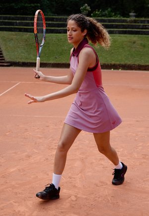 Tennis dress in light pink with a dark pink collar and side panels. Black athletic shoes and white socks, on a clay court background.