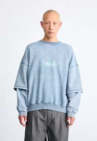 Lys blå oversized hettegenser med ribbestrikket rund hals, med en sentral logo. Stoffet virker mykt og strukturert.