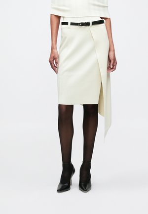 Mulher vestindo uma saia assimétrica off-white com um cinto preto, meias-calças pretas transparentes e sapatos de salto alto pretos contra um fundo branco.