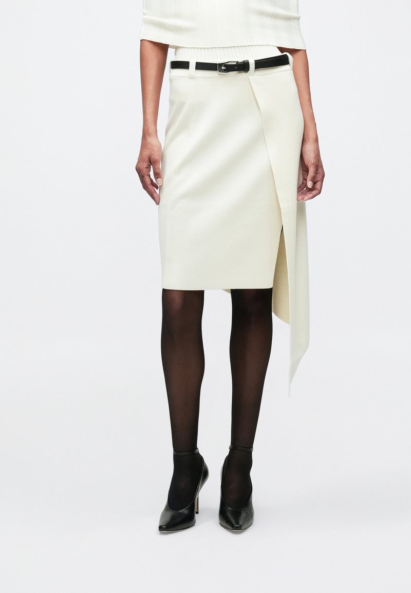 Γυναίκα που φοράει μια εκρού ασύμμετρη φούστα με μαύρη ζώνη, μαύρες διαφανείς καλτσόν και μαύρες ψηλές γόβες με φόντο ένα λευκό φόντο.